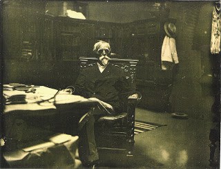 Lew Wallace sits in a chair in his Study next to his desk. Behind him are full bookshelves and a coat rack.