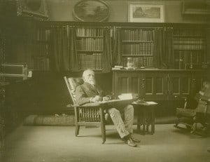 Photograph, black and white. An elderly Lew Wallace sits in a reclining Morris chair inside his Study. Behind him are bookshelves full of books.