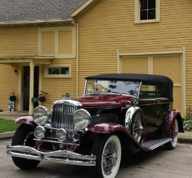 A burgundy 1930 Duesenberg convertible automobile sits in front of the Carriage House