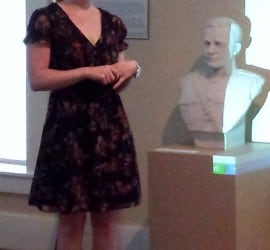A white woman in a dark dress, Allison DePrey, gives a lecture in the Carriage House