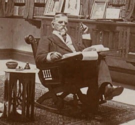 Lew Wallace sits in a rocking chair with a lap desk on his lap. He holds a cigar or pen in his left hand. He is looking at the camera and has gray hair and beard.